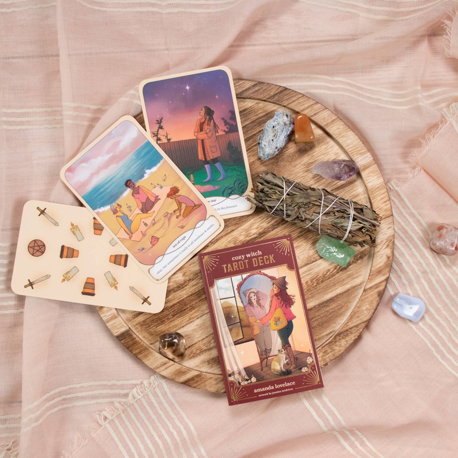 Tarot cards on a wooden board with crystals and smudge sticks on a soft pink fabric background