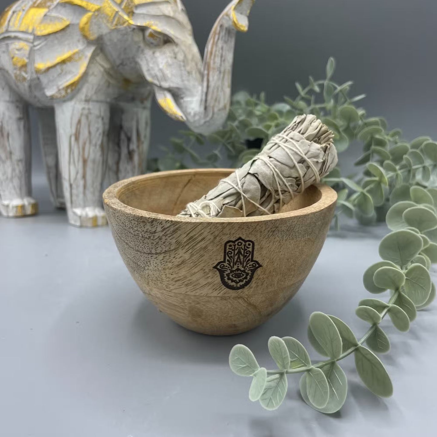 A wooden bowl with Hamsa design containing a sage smudge stick with a decorative background