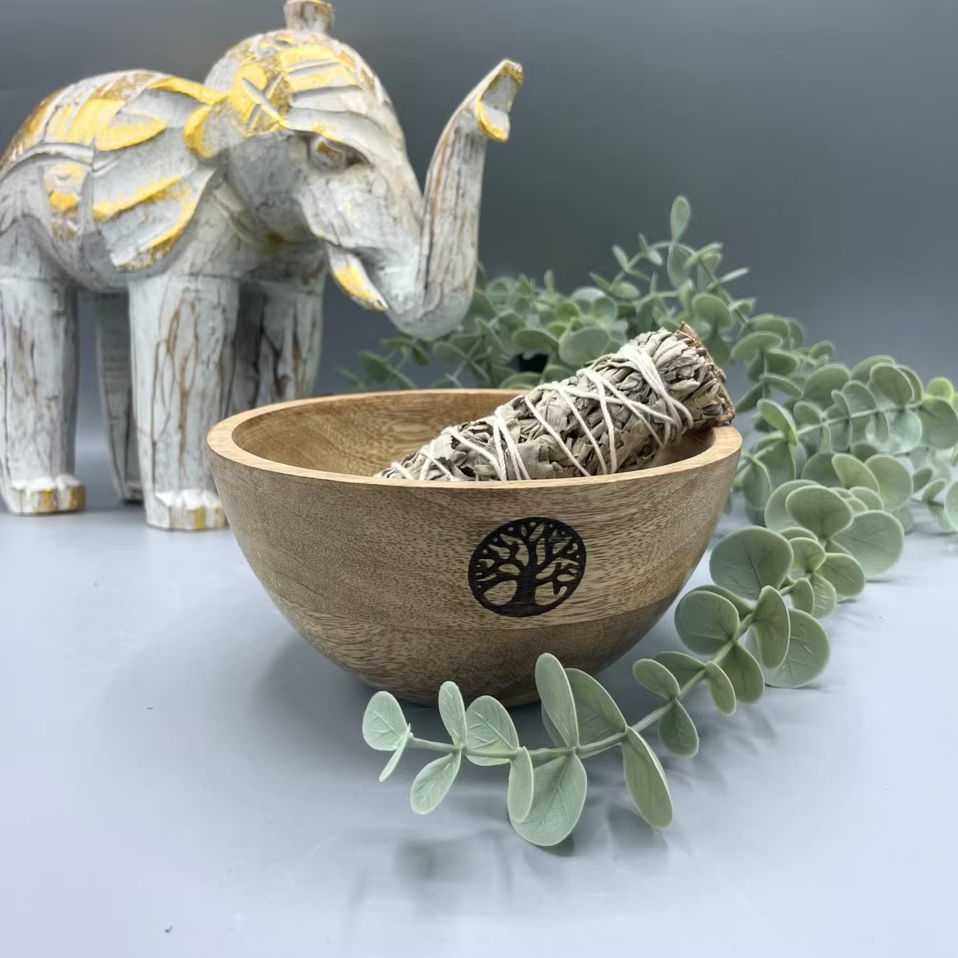 Wooden bowl with sage smudge sticks, eucalyptus leaves, and a decorative elephant on a gray background